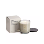 Eine brennende Max Benjamin Italian Apothecary Naturwachskerze in einem Glashalter steht neben ihrer Schachtel und einem dunklen Holzdeckel, alles auf einem weißen Hintergrund.