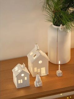 Deux petites figurines de maison blanches en céramique et un bougeoir assorti avec une bougie allumée sont posés sur une surface en bois. À l'arrière-plan se trouve un grand vase blanc avec des branches de sapin vertes.