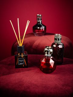 Quatre flacons de parfum élégants, dont un diffuseur à bâtonnets, sont disposés sur deux tabourets ronds et moelleux de couleur bordeaux devant un fond rouge foncé assorti. Les flacons varient en forme et en design et ont un aspect luxueux et sophistiqué.