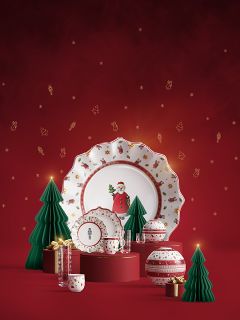 De la vaisselle de fête avec des motifs de Père Noël et de Noël est présentée avec des arbres en papier verts, des boîtes-cadeaux dorées et des ornements rouges devant un fond rouge avec des symboles de fête dorés.