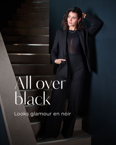 Une femme dans un élégant tailleur pantalon noir est appuyée contre un mur sombre à côté d'un escalier. Le texte sur l'image est : "Tout en noir. On dirait du Glamour en noir". La scène est élégante et moderne.