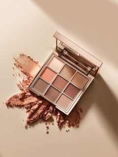 Une palette de maquillage carrée avec neuf fards à paupières dans des tons neutres est ouverte sur une surface beige, où certaines des teintes poudrées sont éparpillées et écrasées. Un miroir est visible dans le couvercle de la palette.