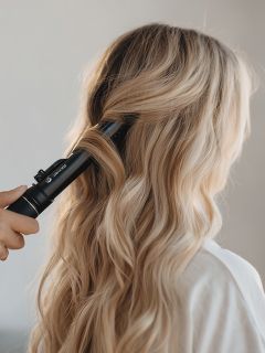 Une personne aux longs cheveux blonds ondulés se fait coiffer par une autre main avec un fer à friser noir. L'accent est mis sur les cheveux et le processus de coiffure.