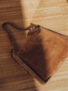 Un sac bandoulière marron en daim avec une longue bandoulière repose sur un plancher en bois baigné d'une lumière chaude du soleil, projetant une ombre douce.