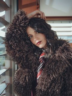 Une femme aux cheveux ondulés et foncés porte un large manteau marron duveteux et une écharpe à motifs, et s'appuie contre une fenêtre. Elle regarde la caméra avec le bras levé, tandis que la lumière du soleil filtre à travers les stores derrière elle.