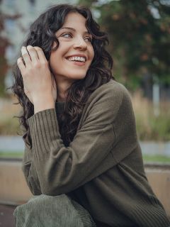 Une femme aux longs cheveux bruns ondulés et portant un pull vert olive est assise à l'extérieur, souriant et regardant sur le côté. Des arbres et des espaces verts sont flous en arrière-plan.