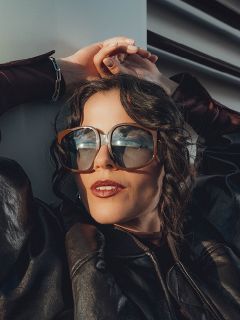 Une femme aux cheveux bruns ondulés porte des lunettes de soleil surdimensionnées et une veste en cuir foncée. Elle est appuyée contre un mur blanc, les mains au-dessus de la tête, et a l'air détendue et élégante.