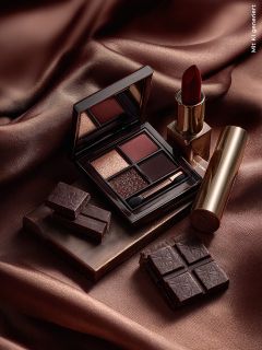 Une palette de fards à paupières, un rouge à lèvres rouge dans un étui doré et des morceaux de chocolat noir sont disposés sur un tissu brun soyeux, formant une composition luxueuse et élégante.