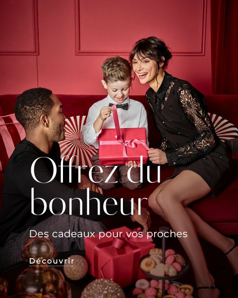 Une famille souriante de trois personnes est assise sur un canapé rouge et échange des boîtes cadeaux roses dans une pièce décorée pour les fêtes, avec des murs rouges et des décorations de Noël. Le texte français est : "Offrez du bonheur. Des cadeaux pour vos proches. Découvrir.