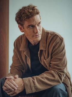 Un homme aux cheveux bouclés châtain clair est assis dans une maison et porte une veste en velours côtelé marron sur une chemise noire. Il regarde pensivement sur le côté et a les mains légèrement croisées. L'arrière-plan est doucement éclairé et neutre.