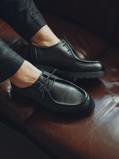 Une personne portant des chaussures à lacets en cuir noir et un pantalon foncé est assise sur un canapé en cuir marron.