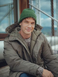 Un homme portant un bonnet en tricot vert, un pull gris et une veste d'hiver vert olive est assis à l'extérieur sur des marches en bois et sourit doucement à la caméra. À l'arrière-plan, on peut voir une clôture métallique et un bâtiment en verre.