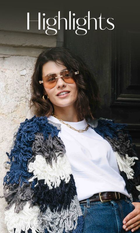 Eine Frau mit gewelltem braunem Haar, die eine Sonnenbrille, einen dicken blau-weißen Strickpullover, ein weißes T-Shirt, Jeans und eine goldene Halskette trägt, steht lächelnd neben einem Gebäude. Über ihr erscheint das Wort "Highlights".