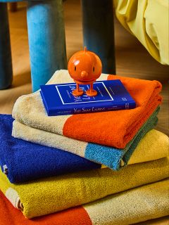 Une pile de serviettes colorées, chacune en blocs audacieux d'orange, de bleu, de jaune et de beige, avec un livre bleu et une petite figurine ronde orange sur le dessus. La scène se déroule dans un intérieur sur un plancher en bois.
