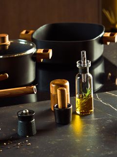 Un plan de travail de cuisine moderne présente des ustensiles de cuisine noirs avec des manches en bois, une bouteille en verre d'huile de romarin, un moulin à poivre, un moulin à sel et un petit mortier avec pilon, le tout sur une surface sombre.