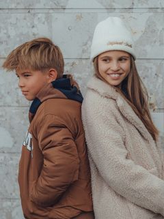 Un garçon et une fille se tiennent dos à dos devant un mur de pierre clair. Le garçon porte une veste marron et regarde sur le côté, tandis que la fille porte un manteau duveteux et un bonnet blanc et sourit en regardant dans la direction opposée du garçon.