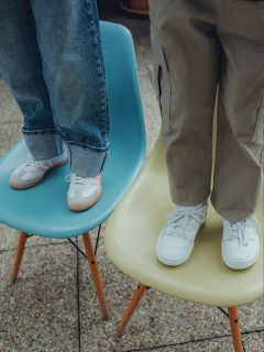 Deux enfants se tiennent debout sur des chaises ; l'un sur une chaise bleue, portant un jean et des baskets à rayures roses, l'autre sur une chaise jaune, portant un pantalon kaki et des baskets blanches. Seules leurs jambes et leurs pieds sont visibles.