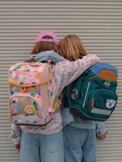 Deux enfants, dos à la caméra, s'embrassent. Ils portent des sacs à dos colorés - l'un de couleur pastel avec des motifs, l'autre vert foncé avec des motifs d'animaux - et des vêtements décontractés, notamment des jeans et une casquette de baseball rose.