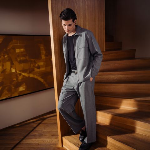Un homme en costume gris avec une chemise noire et des chaussures noires se tient près d'un escalier en bois et s'appuie nonchalamment contre un mur dans un intérieur moderne.