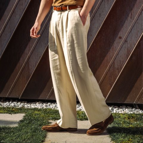 Une personne se tient à l'extérieur, portant un pantalon plissé couleur crème, une ceinture marron, une chemise marron clair et des chaussures marron, la lumière du soleil projetant des ombres sur le sol.