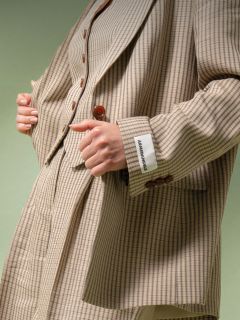 Une personne portant un costume à carreaux beige et marron avec une étiquette blanche portant l'inscription "ABANDONABLE" sur le poignet tient le revers de la veste devant un fond vert.