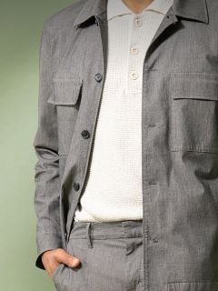 Une personne portant une veste gris clair boutonnée et un pantalon assorti sur un polo blanc texturé se tient debout, une main dans la poche, devant un fond vert atténué.