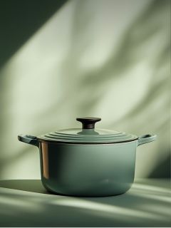 Une cocotte en fonte émaillée verte avec couvercle et bouton noir est posée sur une surface éclairée par une douce lumière naturelle avec des ombres floues en arrière-plan.