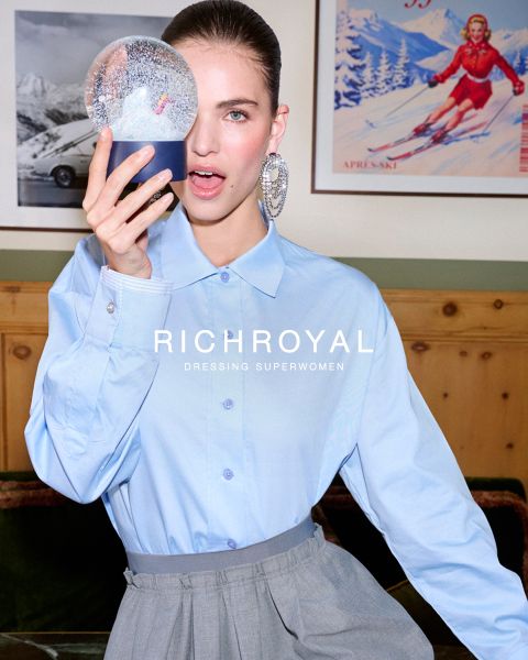 Une femme en chemise bleu clair et pantalon gris tient une boule à neige devant un œil. Elle pose dans une pièce aux murs en bois avec une affiche de ski et porte de grandes boucles d'oreilles. Le texte est "RICHROYAL DRESSING SUPERWOMEN".