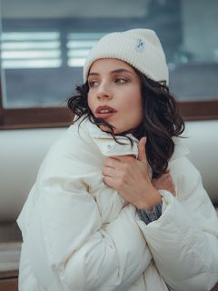 Une femme portant un bonnet en tricot blanc et une doudoune blanche est assise dehors, tenant son manteau fermé et regardant sur le côté avec une expression pensive.