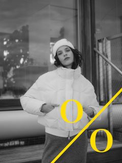 Une personne en doudoune blanche et bonnet pose à l'extérieur près d'un bâtiment ; une ligne diagonale jaune traverse l'image avec deux lettres "O" jaunes de part et d'autre. La photo est en noir et blanc.