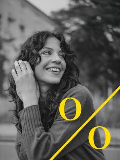 Une femme aux cheveux bouclés sourit et regarde sur le côté. Elle porte un pull et est assise à l'extérieur. Un grand symbole de pourcentage jaune recouvre la photo en noir et blanc.