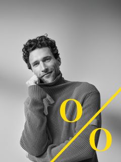 Un homme aux cheveux bouclés et à la barbe porte un gros pull à col roulé et appuie son visage sur sa main en souriant. Un grand symbole de pourcentage jaune est placé en diagonale sur l'image.