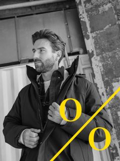 Un homme barbu se tient devant un fond industriel et ajuste sa veste. La photo en noir et blanc montre de gros pourcentages jaunes et une ligne diagonale jaune dans le coin inférieur droit.