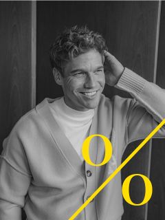 Un homme aux cheveux bouclés, portant un cardigan clair et une chemise blanche, est assis en souriant et tient une main derrière sa tête. L'image en noir et blanc montre deux grands "o" jaunes et une ligne diagonale jaune en bas.