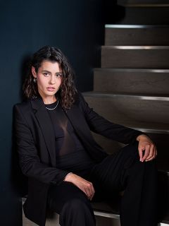 Une femme en tailleur noir est assise sur un escalier en bois devant un mur sombre et regarde l'appareil photo avec assurance, ses cheveux sont coiffés en vagues souples.