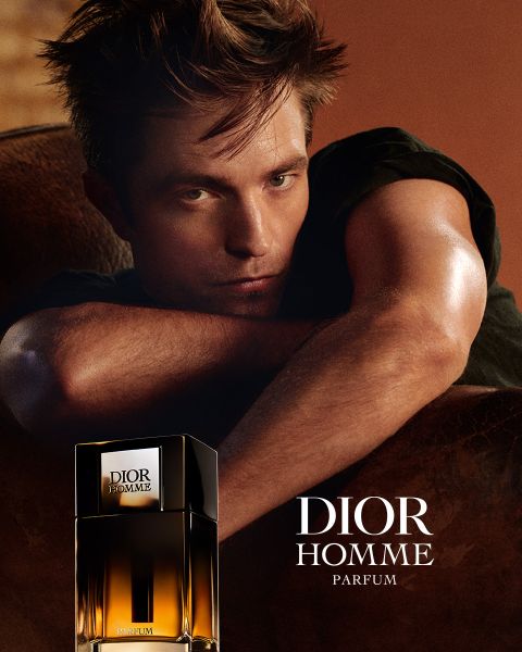 Un homme s'appuie sur une surface marron à côté d'un flacon de Dior Homme Parfum, à côté de lui se trouve le texte "DIOR HOMME PARFUM".