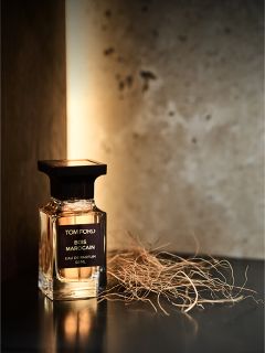 Un flacon du parfum Tom Ford Bois Marocain est posé sur une surface sombre à côté d'herbe séchée, avec un arrière-plan chaud et doucement éclairé.