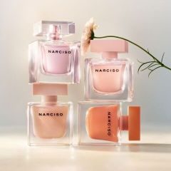 Quatre flacons de parfum de la marque Narciso aux tons pastel sont empilés, et une seule fleur blanche se penche depuis la droite.