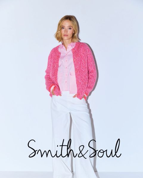 Une femme en chemise rose et cardigan avec un pantalon blanc pose devant un fond blanc. Le texte indique « Smith & Soul » en bas.