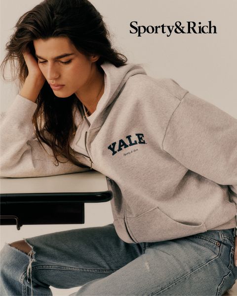 Une personne portant un sweat à capuche gris YALE et un jean déchiré est assise à une table et regarde pensivement vers le bas. Dans le coin supérieur droit, on peut lire "Sporty & Rich".