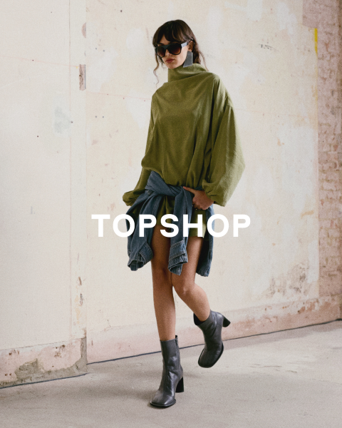 Une femme portant un haut vert surdimensionné, une veste en jean nouée à la taille, des bottes noires et des lunettes de soleil se tient devant un mur usé. Le texte "TOPSHOP" est superposé.
