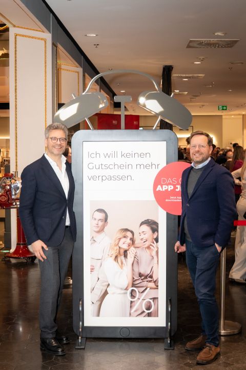 Deux hommes en costume se tiennent à côté d'un grand panneau dans un magasin et font la promotion d'une application avec une photo de trois femmes souriantes et d'un homme sur l'affiche.
