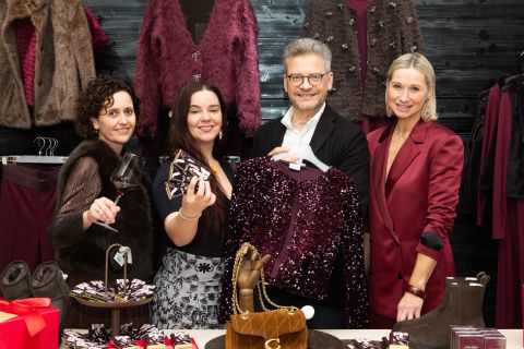 Quatre personnes sourient dans une boutique, tenant une veste à paillettes et des boîtes cadeaux, entourées de robes et d'accessoires bordeaux.