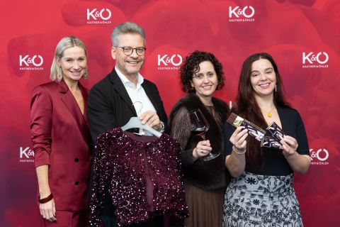 Vier Personen posieren vor einem roten Kastner & Öhler-Hintergrund, halten eine mit Pailletten besetzte Jacke, ein Weinglas und Veranstaltungstickets und lächeln in die Kamera.