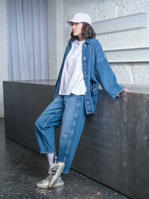 Une personne portant une veste en jean, un t-shirt blanc et une casquette blanche est appuyée contre un comptoir sombre dans un intérieur moderne.