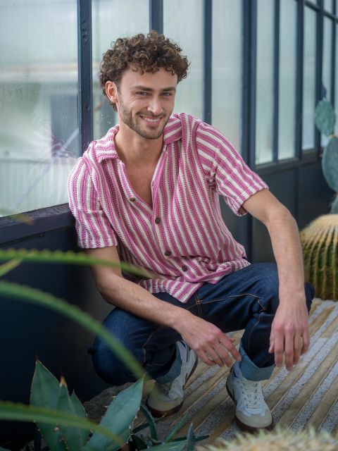 Un homme aux cheveux bouclés, vêtu d'une chemise rayée rose et d'un jean, est accroupi dans une maison près de plantes et sourit à la caméra.