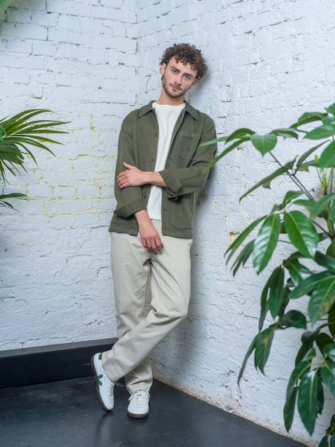 Un homme aux cheveux bouclés, vêtu d'une veste verte, d'une chemise blanche et d'un pantalon beige, est appuyé contre un mur de briques blanches, entouré de plantes vertes.