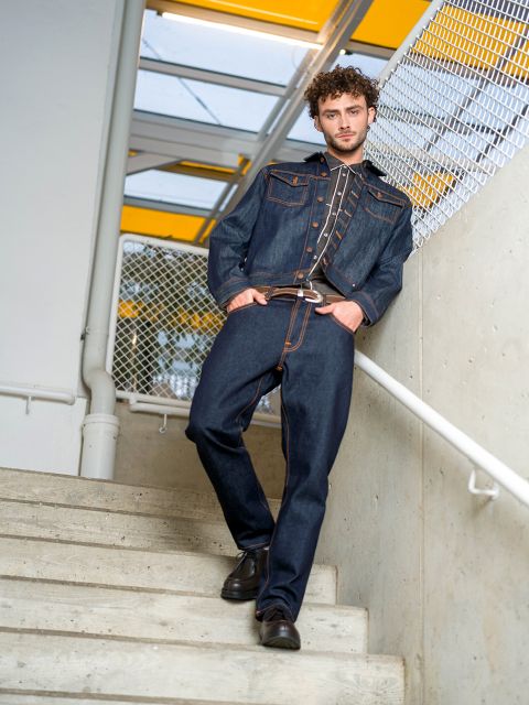 Un homme en veste en jean sombre et en jean se tient sur un escalier, un pied plus haut, et se tient à la rampe, derrière lui un grillage et un lanterneau jaune.