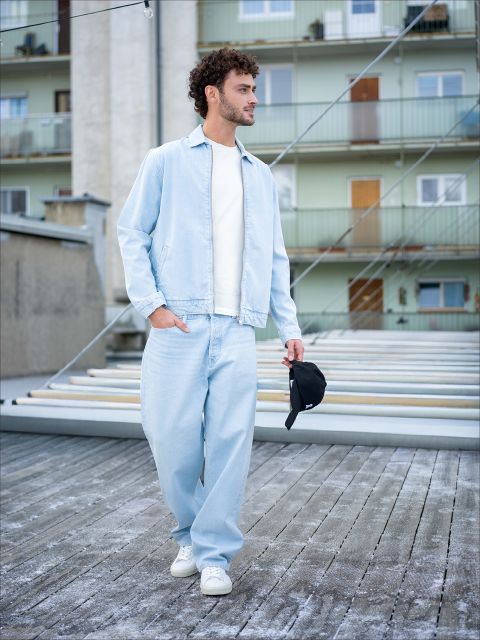 Un homme en jean bleu clair, veste assortie et baskets blanches se promène à l'extérieur et tient un bonnet noir à la main, avec des immeubles résidentiels en arrière-plan.