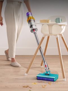 Personne utilisant un aspirateur sans fil pour nettoyer des miettes et des liquides renversés sur un plancher de bois à côté d'une chaise haute et d'une tasse.